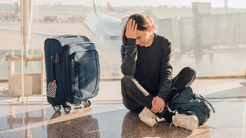 woman sitting on floor of  airport terminal travel bag next to her. Her hand on her head in frustration