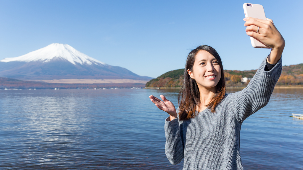 Women holding up phone making video call. In back of her is blue water and Mt Fuji
