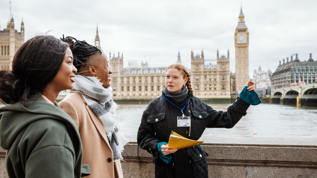 A tour guide gestures while speaking to two people near the River Thames, offering fascinating insights during their tour with the iconic Palace of Westminster and Big Ben visible in the background.
