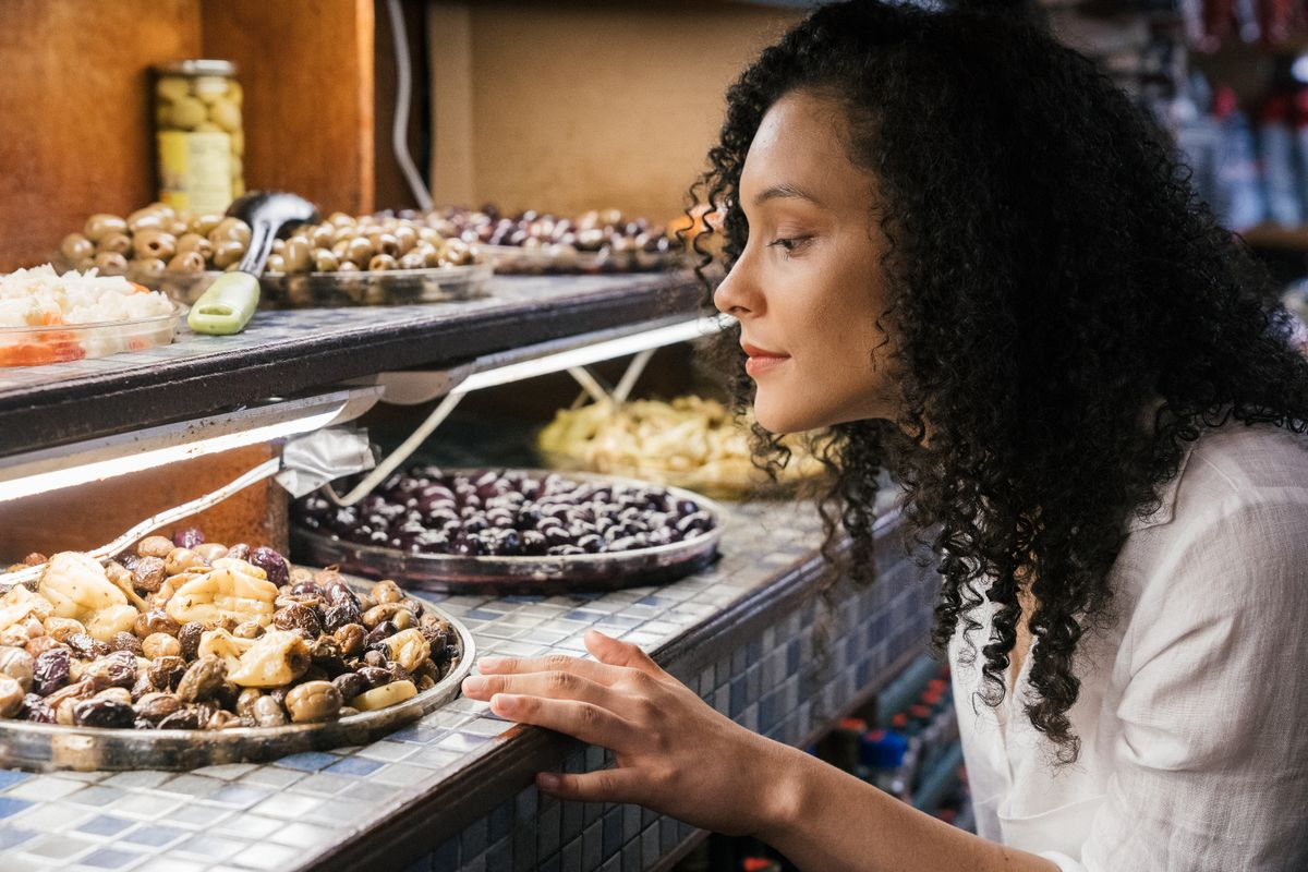 A woman with curly hair examines trays of assorted olives and pickled vegetables displayed on a tiled counter at a market or deli, savoring the sights and flavors on a food tour.